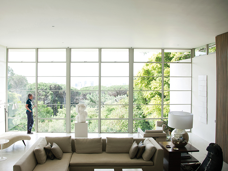Middle aged caucasian man standing in a bright, modern living room gazing out large windows that look out onto a wooded area.