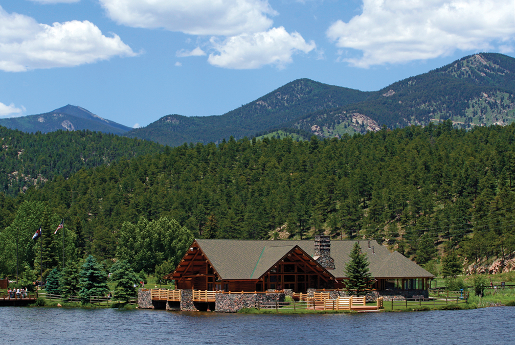 Large wooden house on a lake with evergreen trees and mountains in the distance and a blue sky with white fluffy clouds in Evergreen, Colorado.