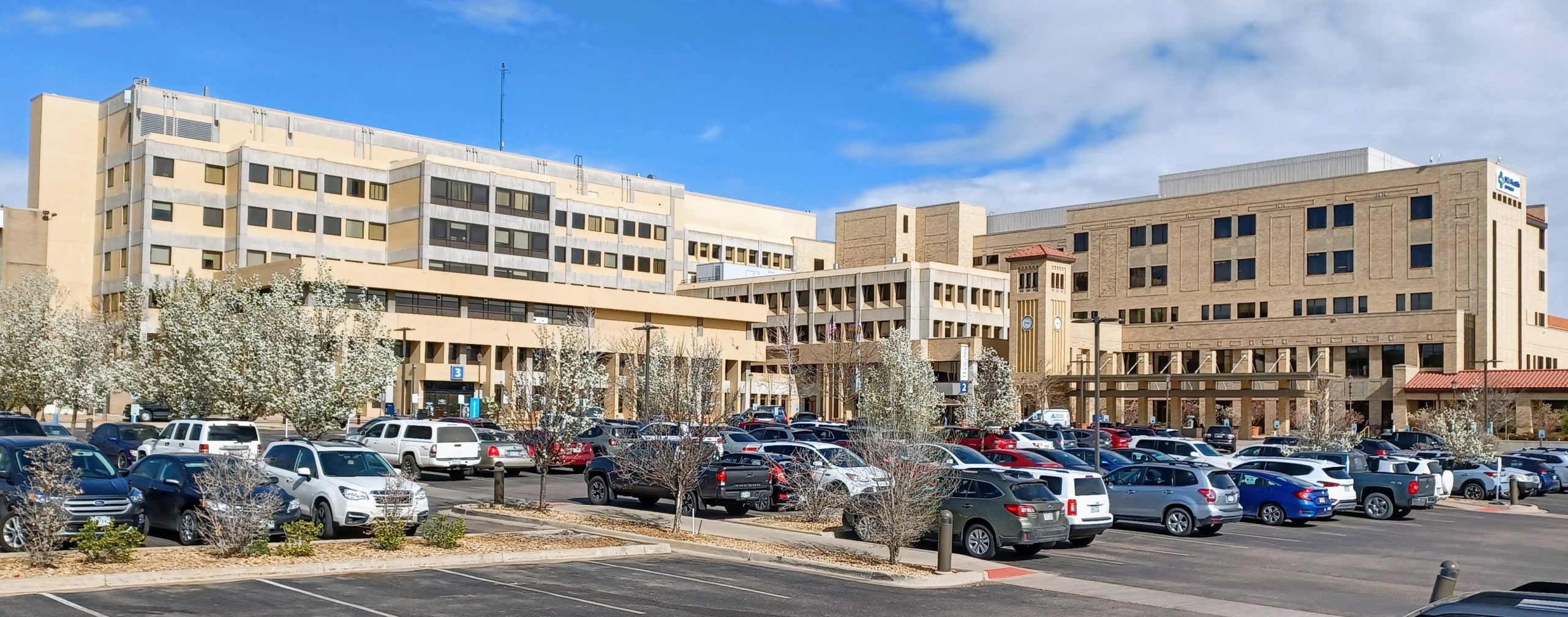Large, light tan hospital building with many cars in the parking lot outside.