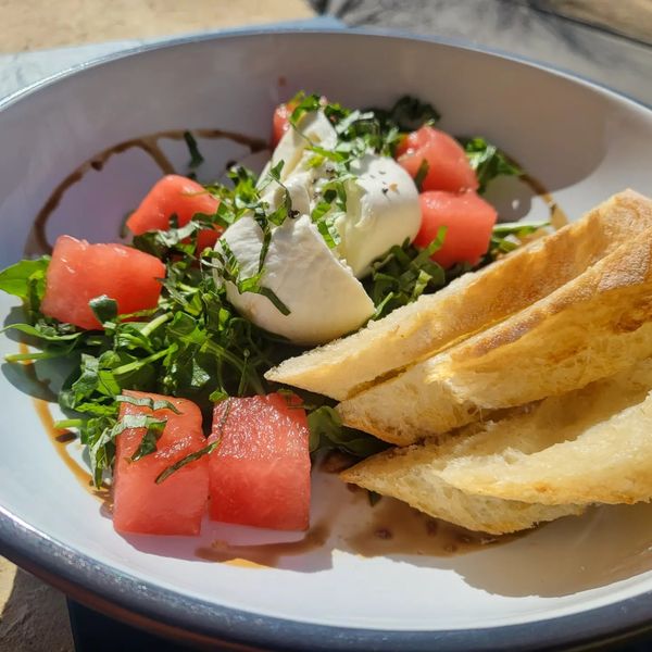Tasty looking plate of bread, salad greens, burrata, and watermelon in natural light.