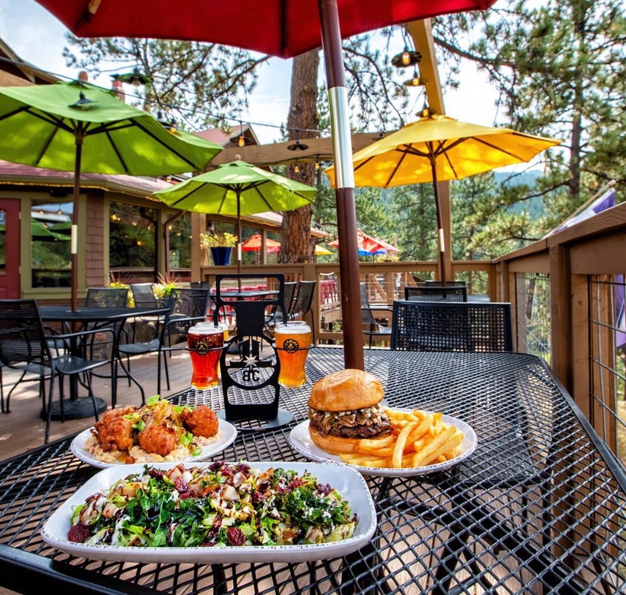 Delicious looking salad, burger and fries, beer flight, and other pub style foods on a table at an outdoor patio restaurant with colorful umbrellas surrounded by evergreen trees.