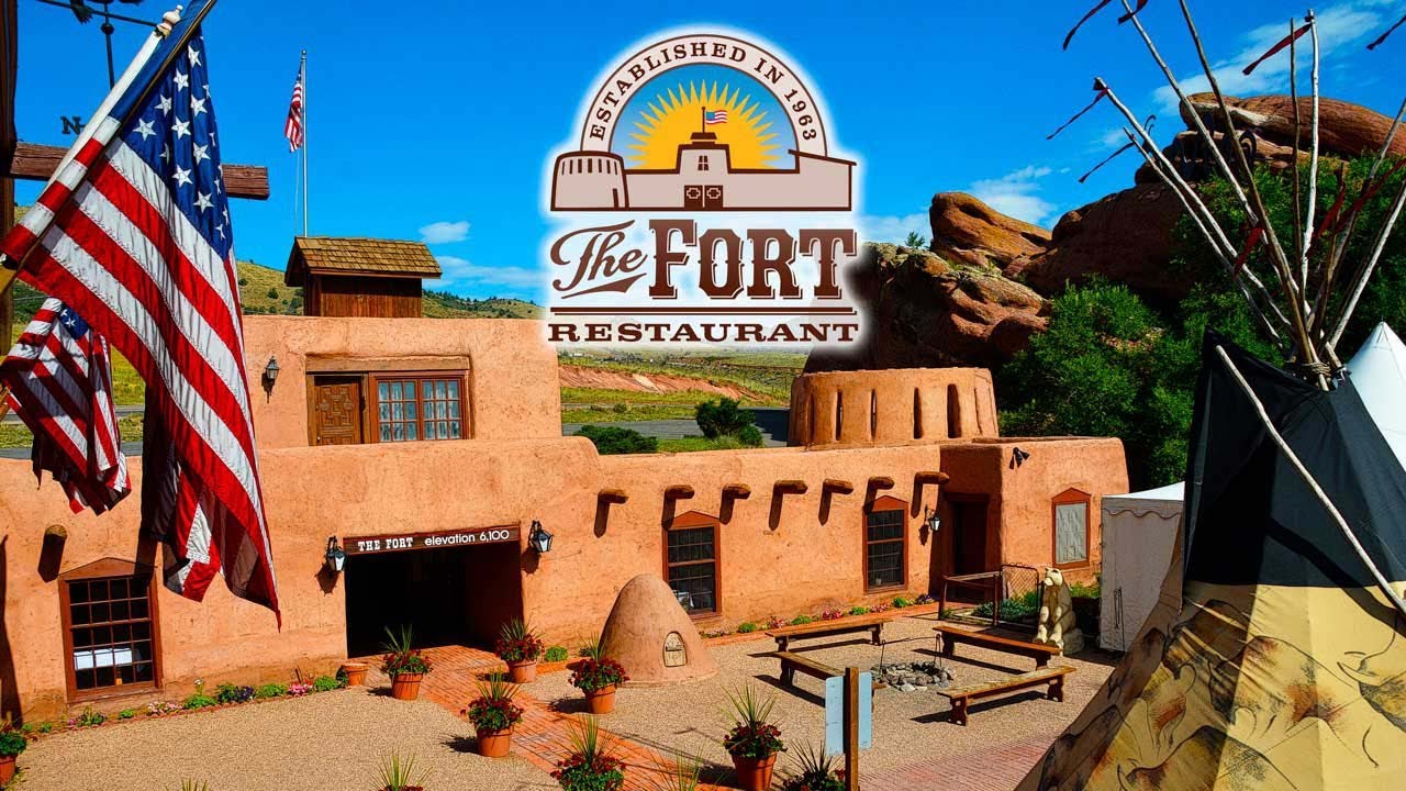 Exterior of an Adobe style building with American flags and the top of a teepee. Overlaid with the logo for the Fort Restaurant in Morrison, Colorado.