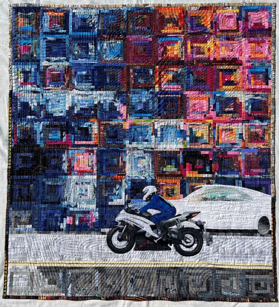 Colorful quilt with person on motorcycle and a white car.