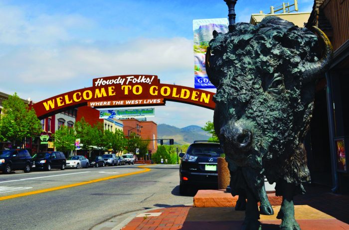 Statue of a Buffalo on a car-lined street in downtown Golden with a red arch that goes over the street that says "Howdy Folks! Welcome to Golden, Where the West Lives"