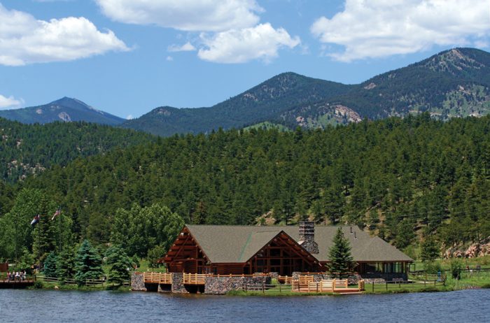 Large wooden house on a lake with evergreen trees and mountains in the distance and a blue sky with white fluffy clouds in Evergreen, Colorado.