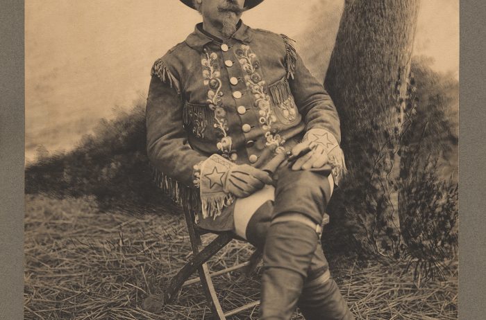 An antique, sepia toned photo of Buffalo Bill sitting with legs crossed. He is wearing boots, gloves, and a cowboy hat.