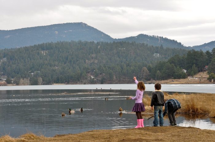 Three kids standing in front of a lake with mountains in the distance and ducks in the water.