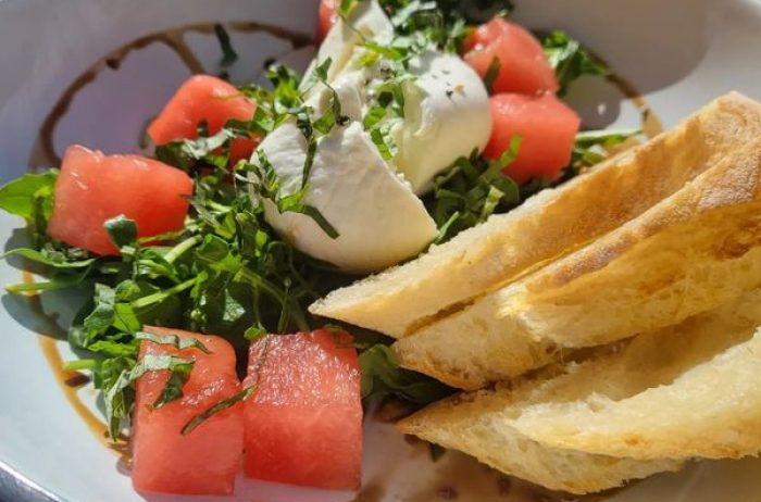 Tasty looking plate of bread, salad greens, burrata, and watermelon in natural light.