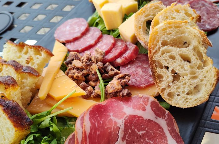 Close up of a charcuterie plate on a black board with bread, meat, cheese, greens, and nuts on a table outdoors.