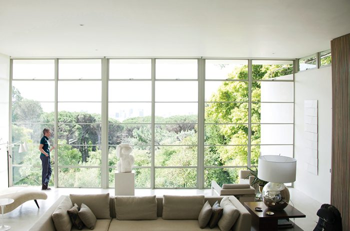 Middle aged caucasian man standing in a bright, modern living room gazing out large windows that look out onto a wooded area.