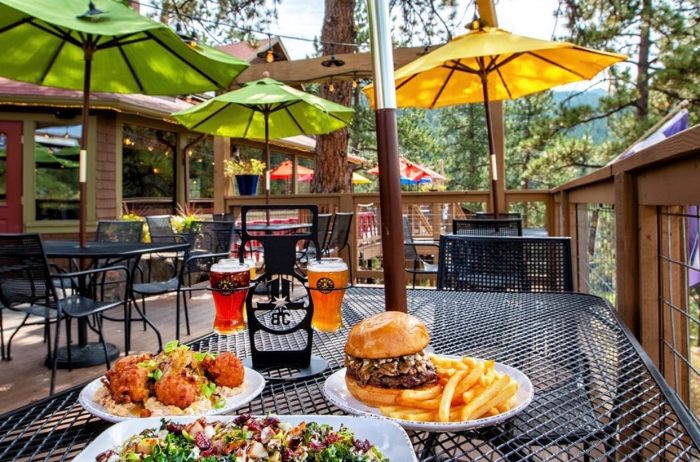 Delicious looking salad, burger and fries, beer flight, and other pub style foods on a table at an outdoor patio restaurant with colorful umbrellas surrounded by evergreen trees.