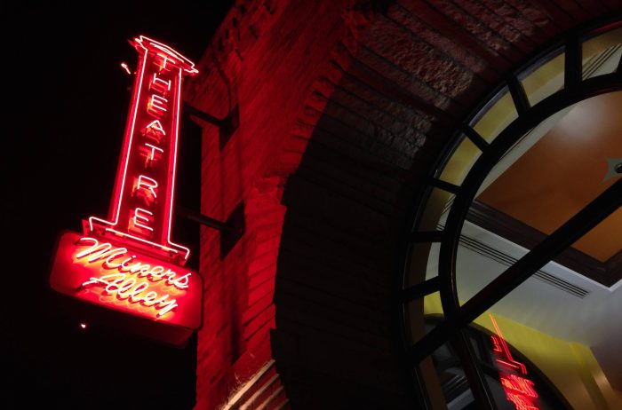 Sign for Miners Alley Theater lit up in red on the exterior of a brick building at night.